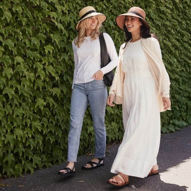 Two women walking outdoors with a green hedge background wearing the Solbari Provence hat.